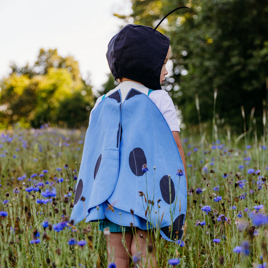 A child standing in a wildflower field, viewed from behind, wearing blue butterfly costume wings made from linen and cotton, paired with a black antennae hat