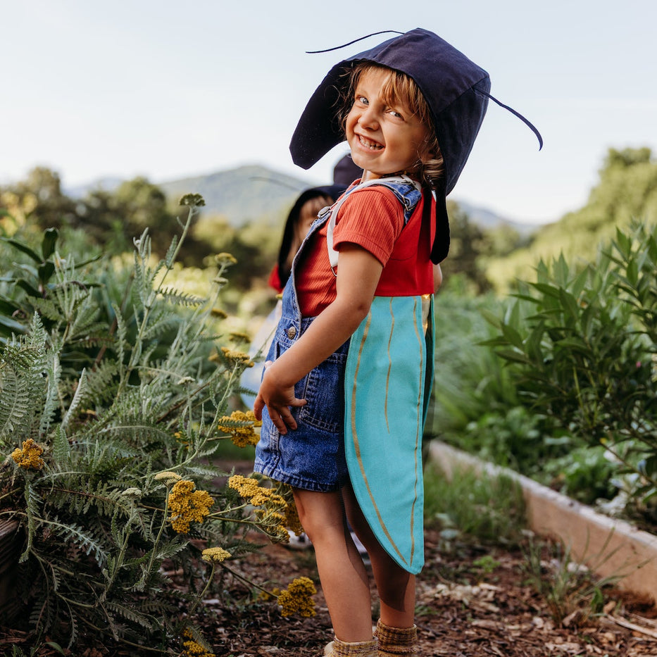 A young child standing in a garden smiling as they play in bug costume wings and a matching antennae hat. The costume is made out of natural fibers. 