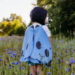 A child standing in a wildflower field, viewed from behind, wearing blue butterfly costume wings made from linen and cotton, paired with a black antennae hat