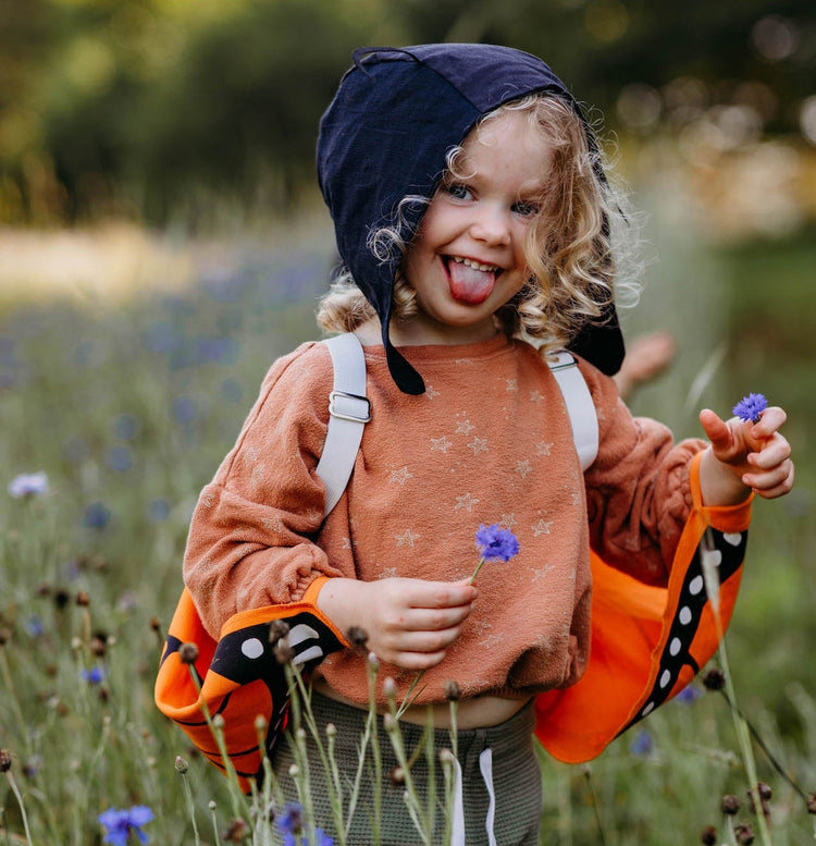 A child playing in a field of purple wildflowers wearing natural fiber butterfly costume wings for kinds. The child also wears a matching natural fiber antennae hat.
